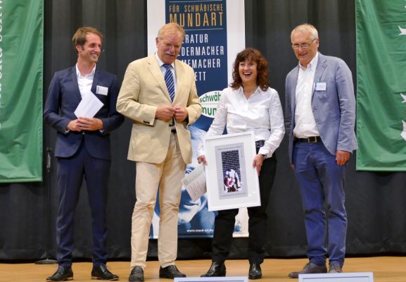 Marion Kinzig erhält auf der Bühne den Sebastian‑Blau‑Preis 2018 und nimmt die Urkunde vom Vorstand des Vereins Mundart entgegen.